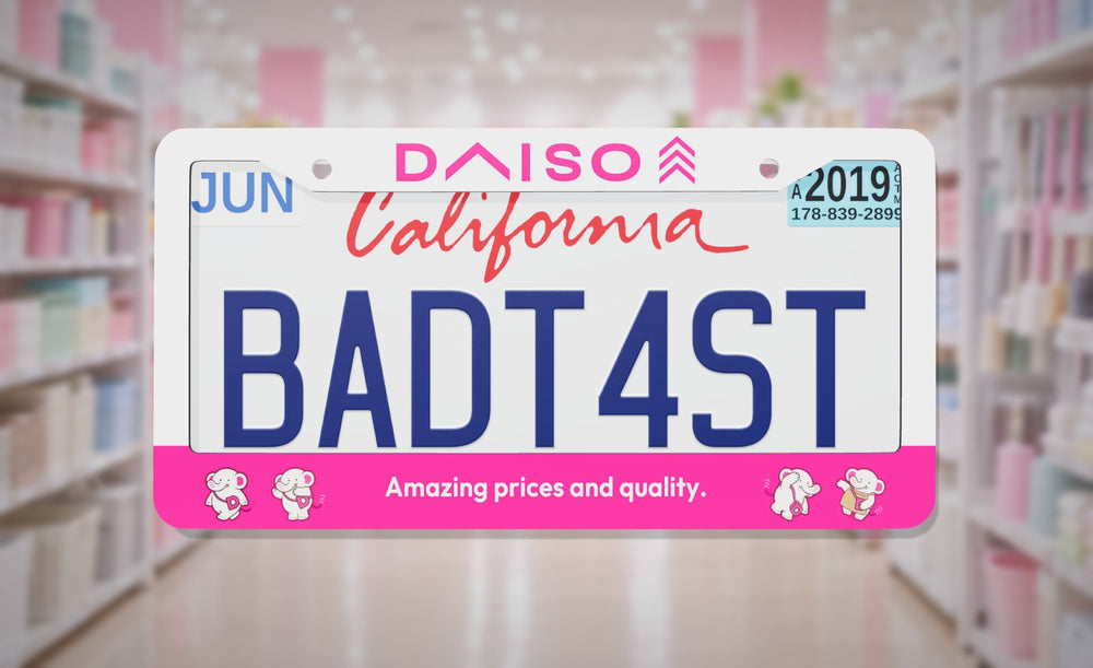 Daiso license plate frame in white and pink with the Daiso logo and "Amazing prices and quality.", front view in a bright pink and white variety store aisle