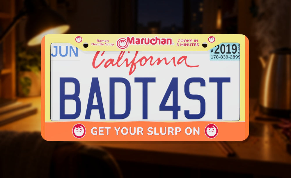 Maruchan license plate frame in orange with the Maruchan logo and Get Your Slurp On, front view on a late-night desk background