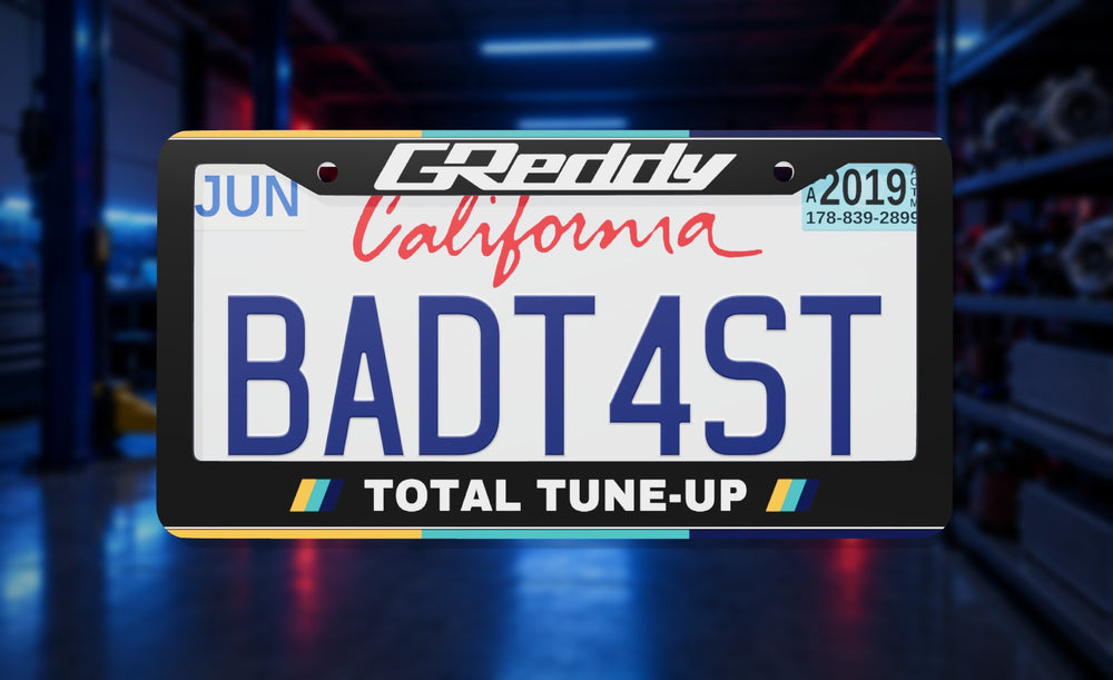 GReddy license plate frame in black with the GReddy logo and Total Tune-Up, front view on a neon-lit tuning garage background