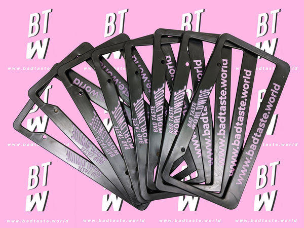 Stack of black Bad Taste Worldwide license plate frames on a pink background
